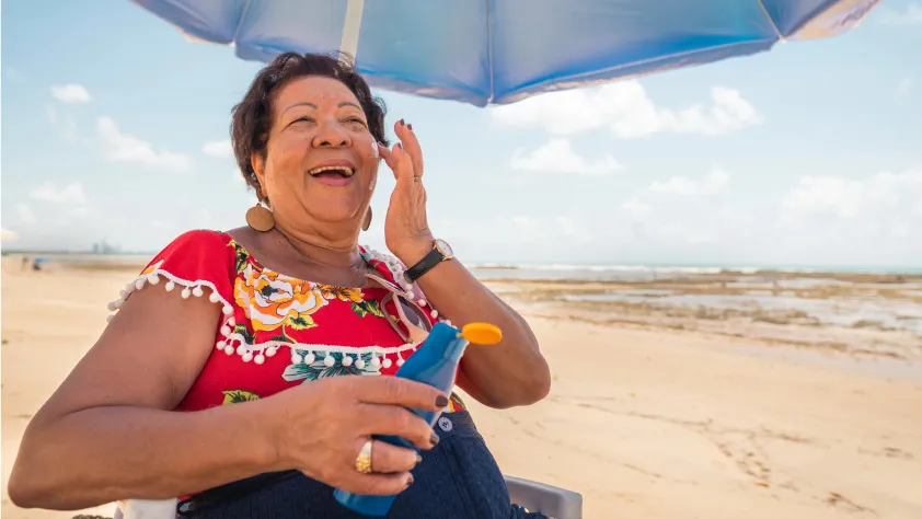 Person sitting at the beach underneath umbrella applying sunscreen to face