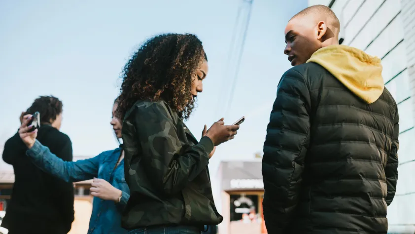 One person taking picture on phone in background while two people are standing in front and one is scrolling through phone