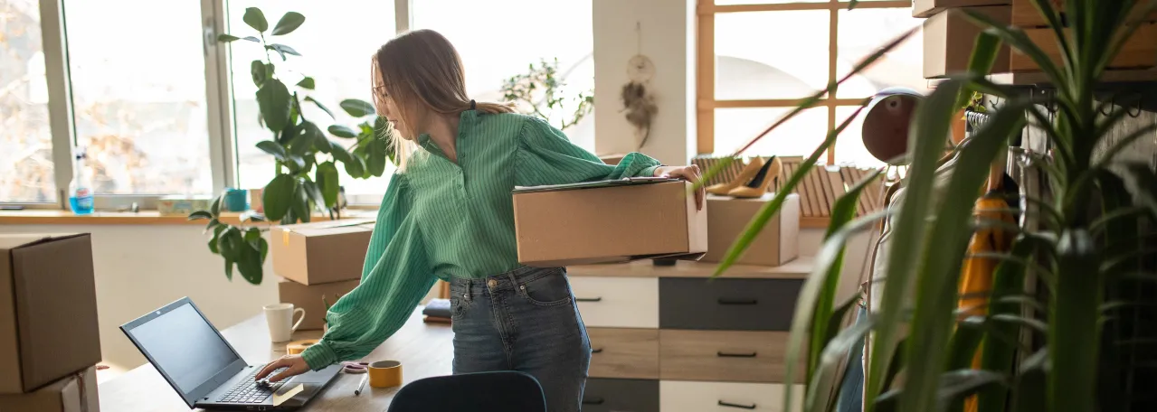Person in office standing at desk while holding a box with one arm and typing on laptop with the other