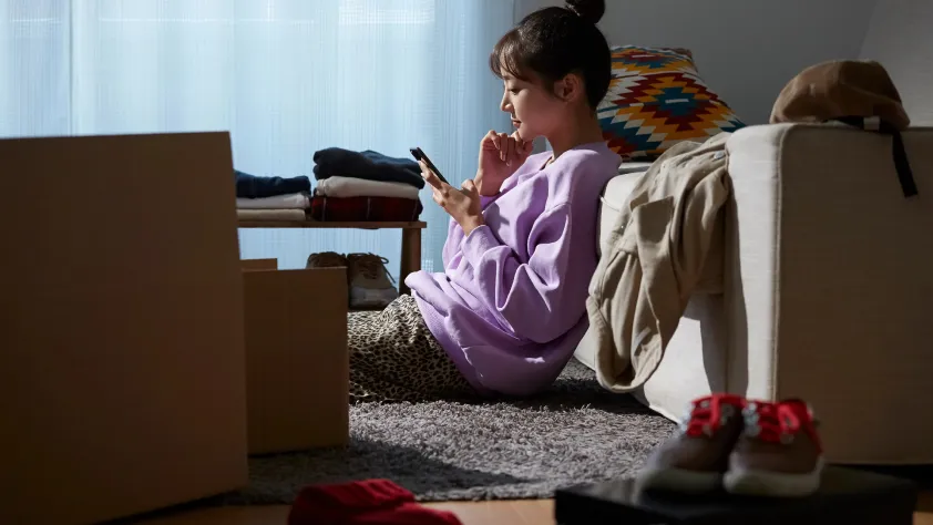 Person sitting on floor in the middle of unpacking, leaning against couch while scrolling on phone