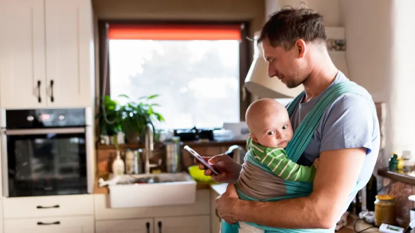 Person standing in kitchen while holding a baby and checking phone