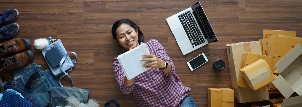 Aerial view of person laying on floor and taking notes while surrounded by laptop, phone, boxes, and clothes