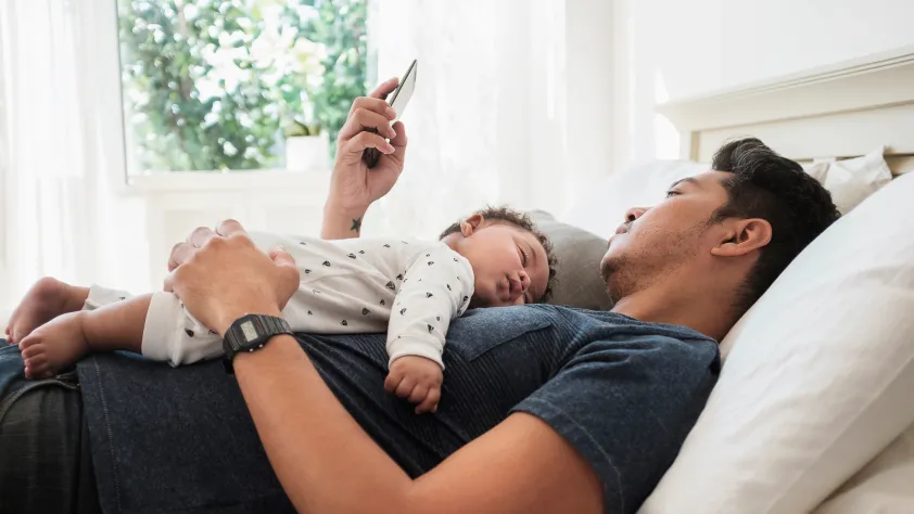 Pessoa deitada na cama usando o smartphone e com um bebê dormindo sobre o peito