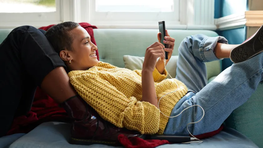 Pessoa deitada no sofá encostada nas pernas de outra pessoa e sorrindo enquanto olha o celular