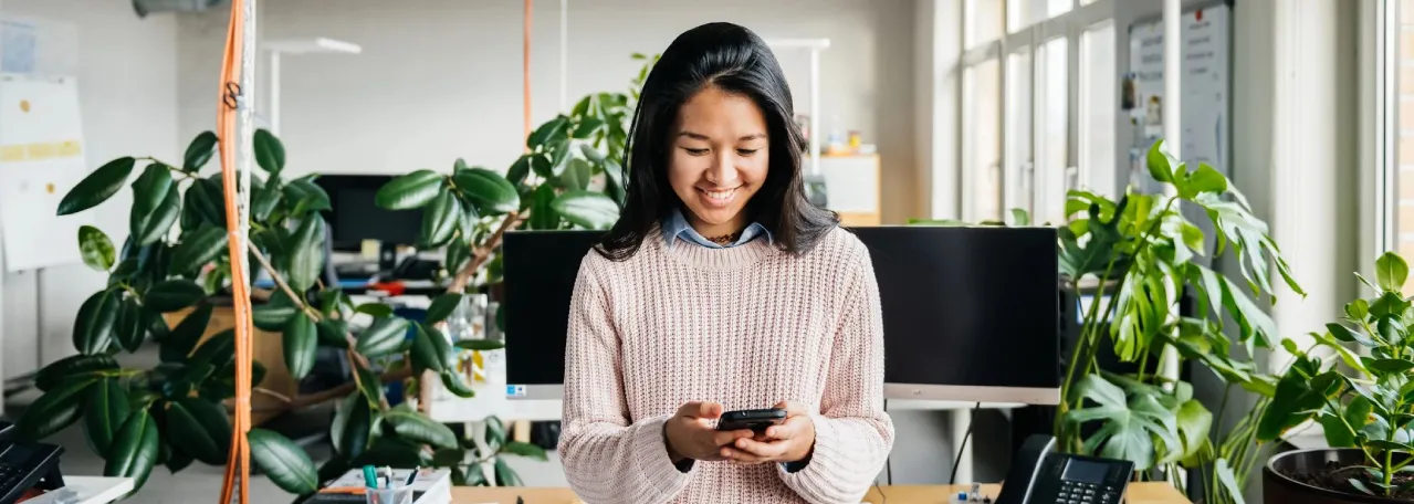 Pessoa em pé no escritório sorrindo enquanto olha para o celular