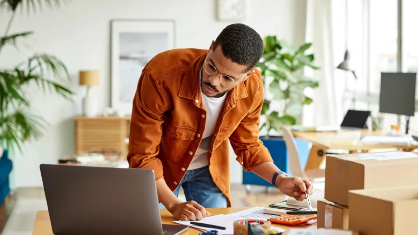 Person standing at desk in home office reading on his laptop while taking notes