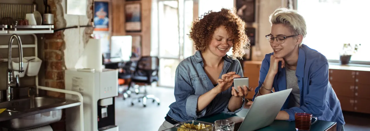 Two people standing in office kitchen while smiling at phone