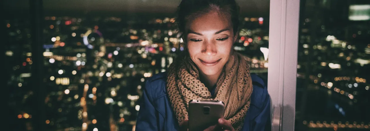 Person standing in front of window while looking at phone with the night skyline in the background