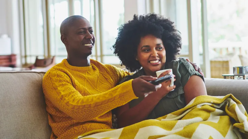 Two people sitting on couch smiling, while flipping through TV channels