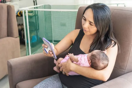 A woman looking at her phone and holding a baby while sitting on a sofa