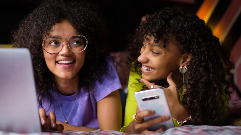 Two people laying on bed while smiling and working on their laptop and phone