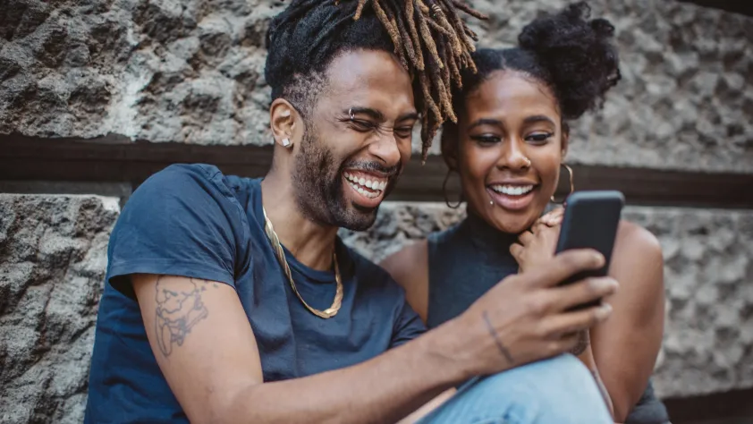 Two people sitting against a wall while laughing at an ad they are seeing on the phone
