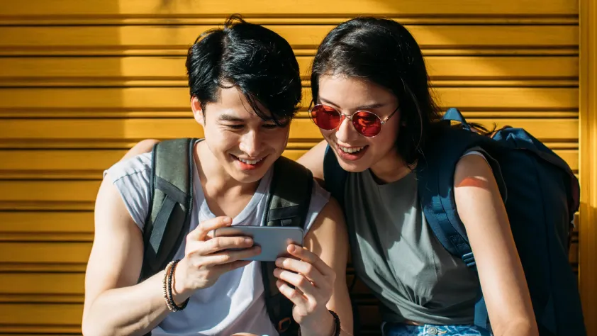 Two people wearing backpacks smiling while watching an ad on phone