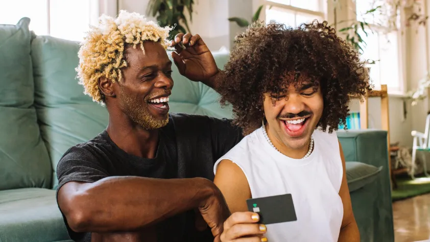 Two people laughing while sitting against couch and one is holding a credit card