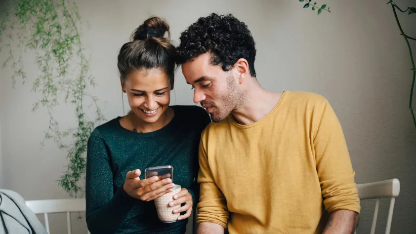 Two people sitting on bench indoors and one is holding a coffee and they are both looking at a business message via WhatsApp