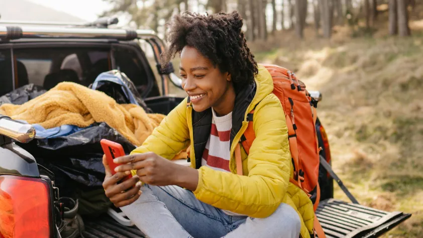 Person sitting on the back of a truck with camping gear in the woods while smiling on a video call