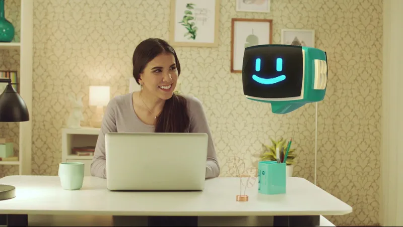 Seorang wanita duduk di depan komputer laptop dengan kepala robot yang tersenyum melayang di udara di sampingnya.