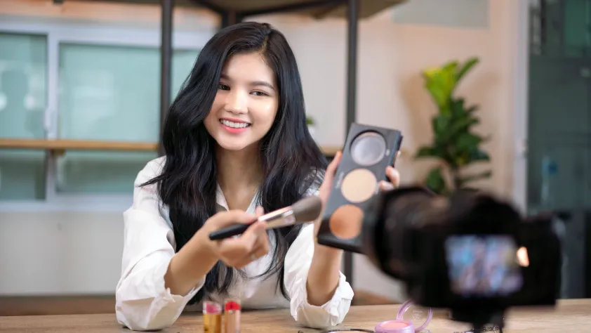 Person smiling and showing a makeup palette and brush while filming a makeup tutorial
