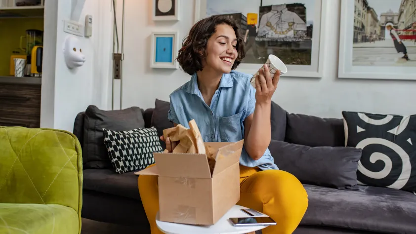 Person sitting in living room on a gray couch while opening a package containing a ceramic cup