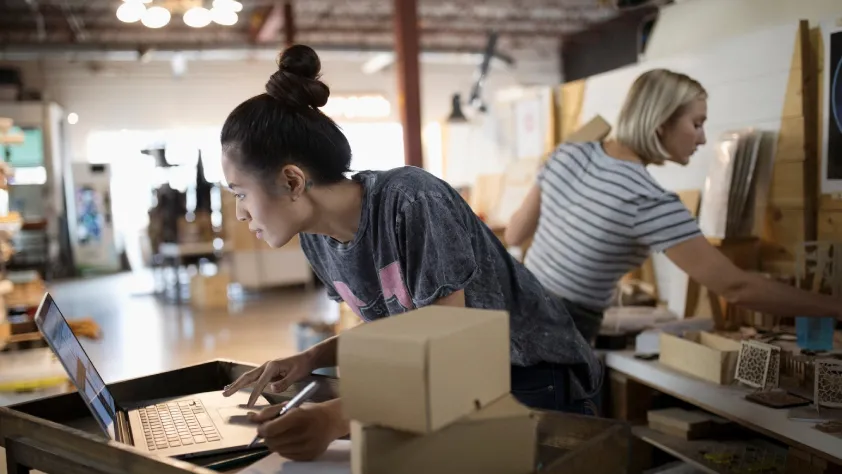 Two people working in an art studio. One is putting together an order and the other person is writing down an online order from a laptop.