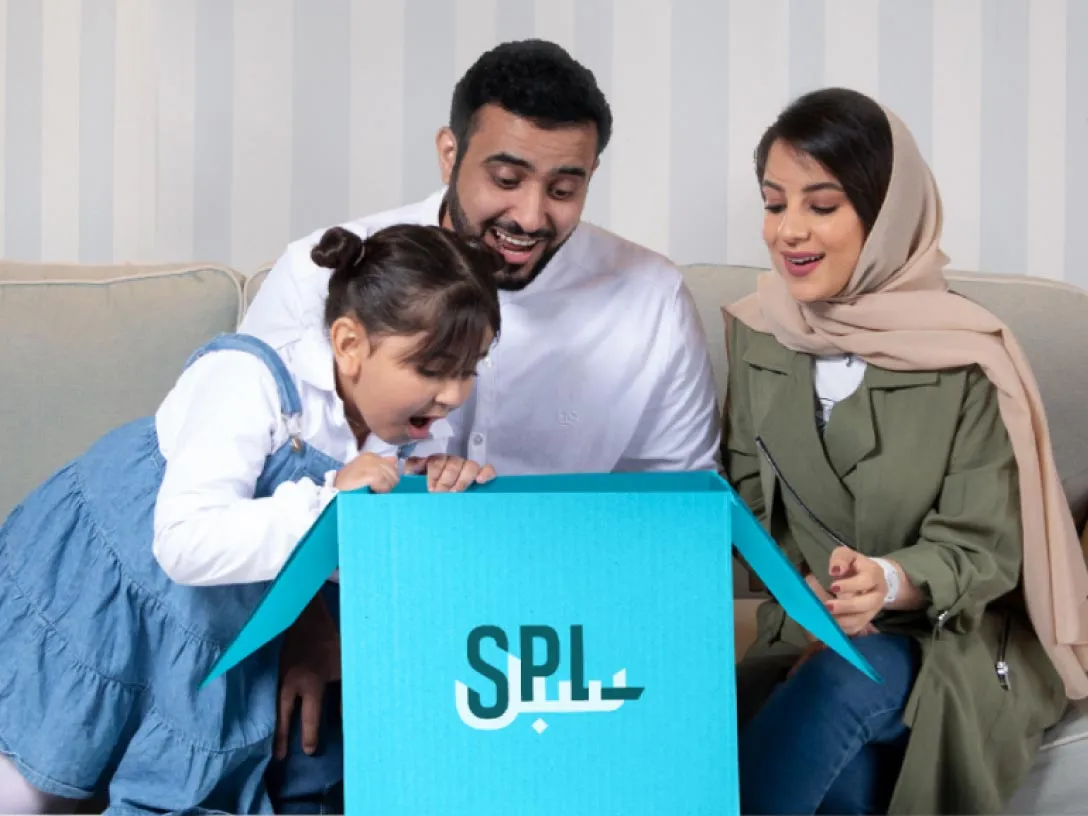 Pais sentados em um sofá enquanto a filha parece surpresa com um pacote aberto da SPL.