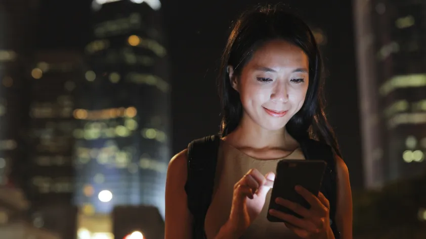 Pessoa andando pela cidade à noite enquanto olha para o smartphone. A luz do telefone está brilhando forte em seu rosto.