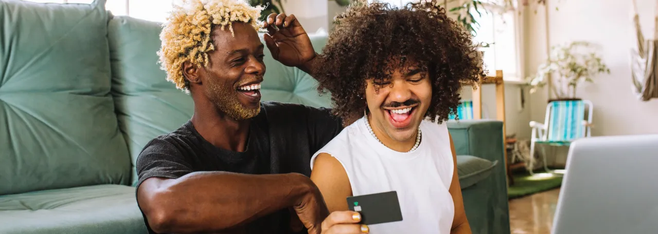 Two people laughing while sitting against couch and one is holding a credit card