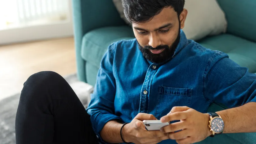 Pessoa sentada no chão encostada em um sofá azul enquanto conversa pelo WhatsApp no smartphone