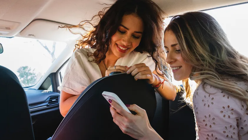Two people in car. One is turning around from the passenger seat to look at the phone of the person sitting in the backseat