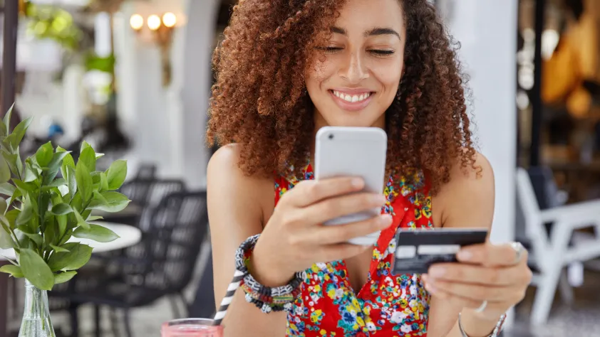 Person sitting at an outdoor cafe, smiling, while holding credit card in one hand and making a purchase on phone in the other hand