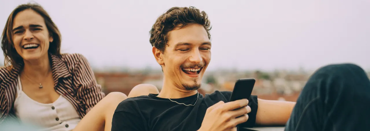 Two people sitting outside laughing while the person in front is scrolling on their phone