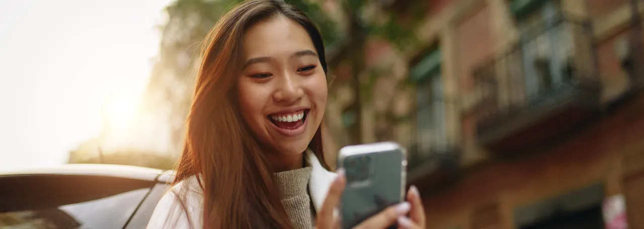 Person walking down the street with sun shining in the background while smiling and making a purchase on phone