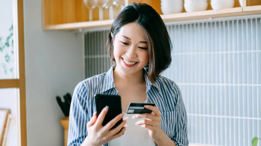 Person standing in kitchen, smiling, while holding credit card to make a purchase on phone