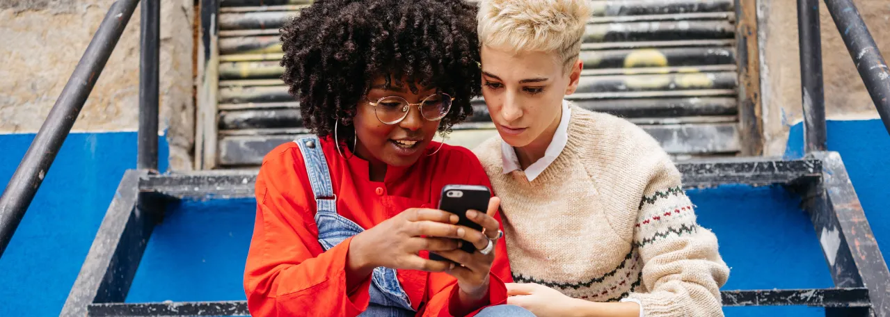 Duas pessoas sentadas nos degraus de uma escada ao ar livre. Uma pessoa mostra à outra uma mensagem de uma empresa no WhatsApp em seu smartphone