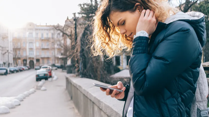 Person leaning against a concrete wall outside on the street with snow on the ground. They are reading a WhatsApp message on phone.