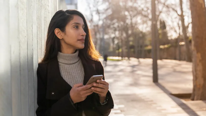 Person leaning against wall in outdoor park while holding phone and looking into the distance