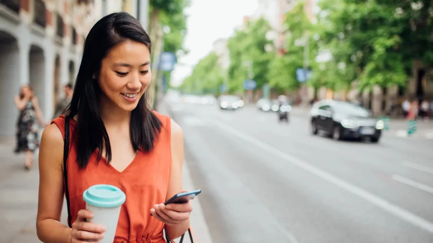 Person smiling and walking down the street while holding a cup of coffee and engaging with a WhatsApp message