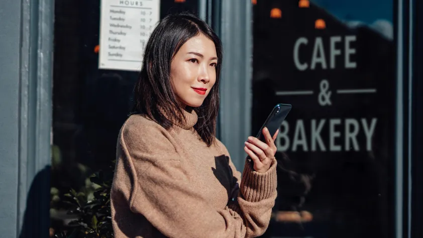 Person standing outside of cafe and bakery, wearing a sweater and red lipstick, while holding phone