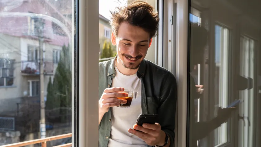 Person sitting in windowsill while smiling and holding an espresso and reading a business message via WhatsApp on phone