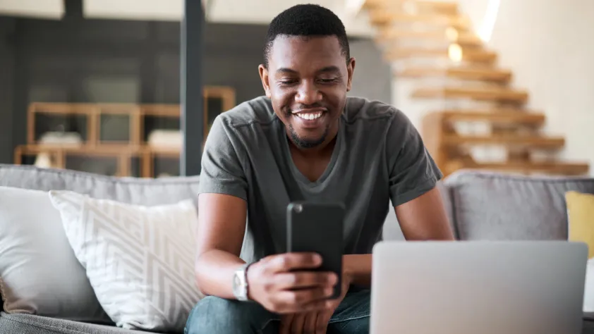 Person smiling and sitting on living room couch in front of laptop while reading a WhatsApp marketing message on phone