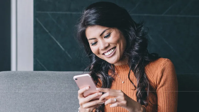 Person sitting on gray couch in office, wearing an orange long sleeve shirt, while smiling and reading a WhatsApp business message
