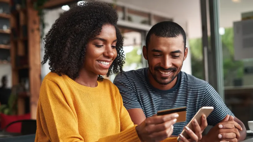 Two people sitting next to each other at a table, both smiling. One is holding a credit card and making a purchase on their phone through WhatsApp.