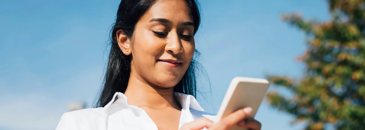 Person smiling at phone, wearing a white button down shirt, in front of a blue sky background with a tree