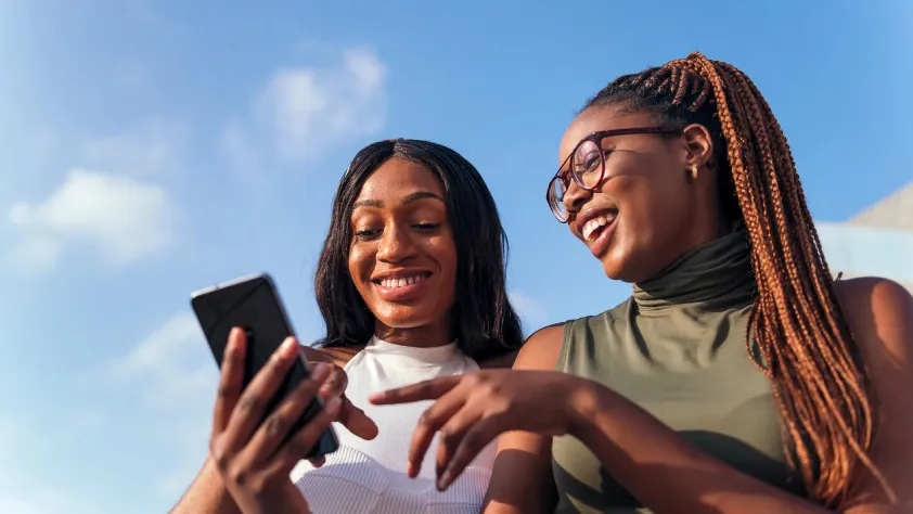 Two people sitting with blue sky in background, while laughing and looking at one of their phones