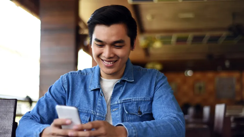 Person sitting in cafe, wearing a jean jacket and smiling while reading a WhatsApp message on phone