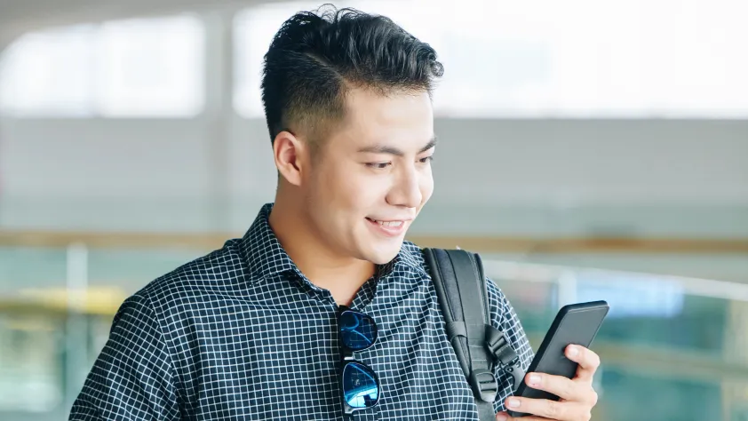 Person wearing a button down shirt with sunglasses hanging on the collar. They are walking through the office while smiling and looking at an ad on phone.
