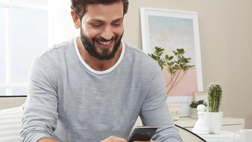 Person sitting in living room with white furniture. They are wearing a gray long sleeve shirt and smiling while reading a personalized WhatsApp message from Tata CLiQ.