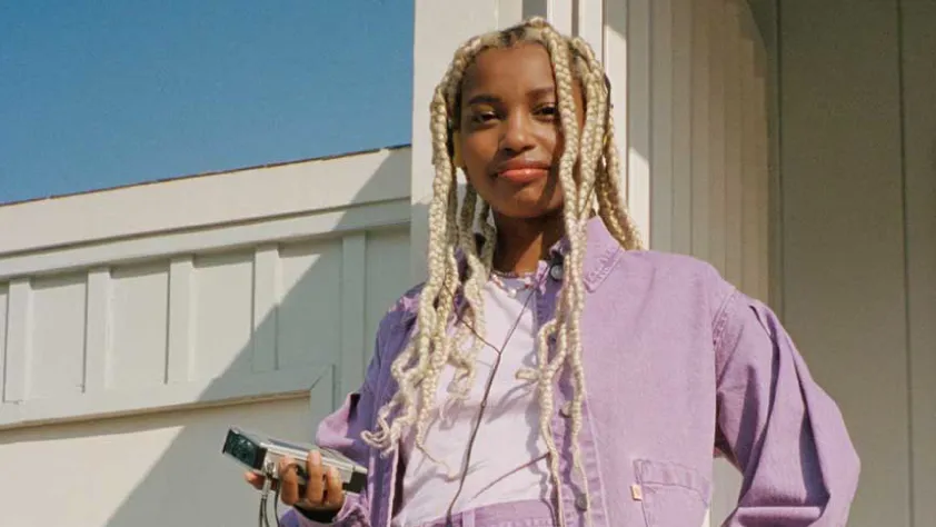 Person standing against white wall and wearing a purple denim jacket. They have headphones in that are plugged into a cassette player.