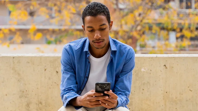 Person sitting on a bench with a tree in the background. They are wearing a white t-shirt with a blue denim long sleeve shirt while opening a WhatsApp message on phone.
