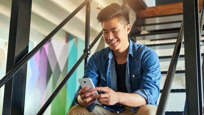Person sitting on office stairs, wearing a denim shirt and khakis. They are smiling and submitting a customer inquiry through WhatsApp.